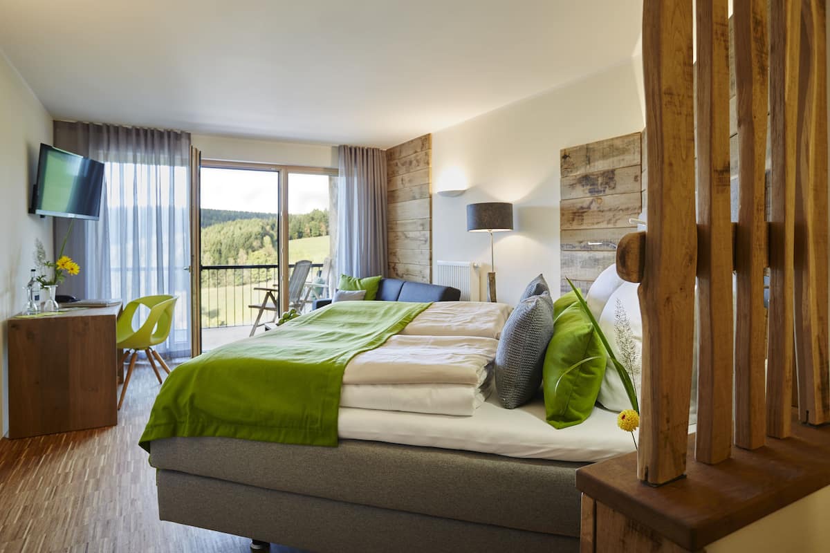 A modern hotel room with a large bed, a television, a desk, and a balcony with a view of the countryside.