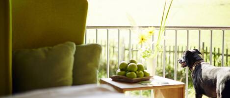 Una habitación con una cama, un sofá verde, una pequeña mesa con un frutero y un jarrón de flores, y un perro junto a un balcón con vistas a un campo verde.