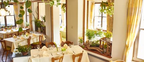 Un restaurante con varias mesas dispuestas para comer, sillas de madera y amplias ventanas con cortinas.