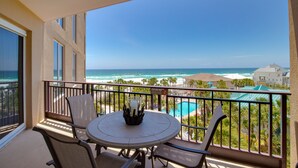 A balcony with a table and chairs, overlooking a pool and ocean view. - Updated Beachfront Condo - Beach View, Balcony, Pool, Tiki Bar, Hot Tub, Tram (Miramar Beach)