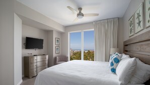 A bedroom with a large bed, a ceiling fan, a TV, a dresser, and a view of the ocean through the window. - Updated Beachfront Condo - Beach View, Balcony, Pool, Tiki Bar, Hot Tub, Tram (Miramar Beach)