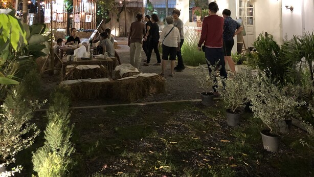Un área de comedor al aire libre con mesas y sillas, rodeada de plantas en macetas y árboles.