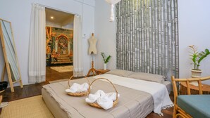 Habitación de hotel con una cama, dos cestas de mimbre con toallas, una silla de madera, una mesita, una planta en maceta y un panel decorativo en la pared.