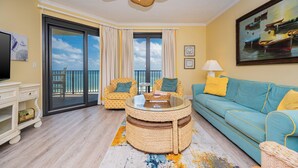 A living room with a teal sofa, yellow patterned chairs, a glass coffee table, and a balcony with a view. - Lovely 3BR Gulf Front Condo - Phoenix VII 71205 (Orange Beach)