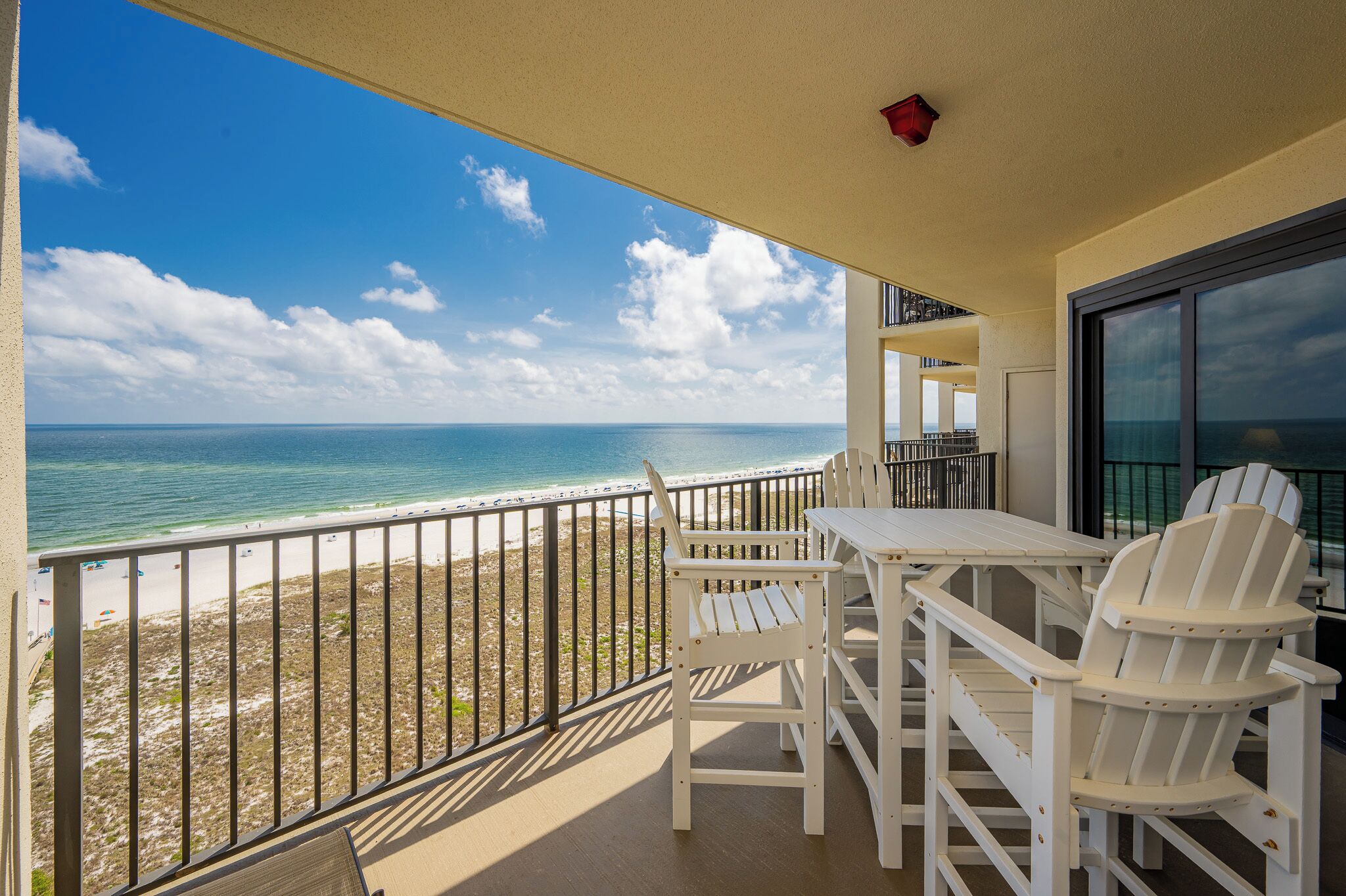 A balcony with a view of the beach and ocean, white chairs, and a table.