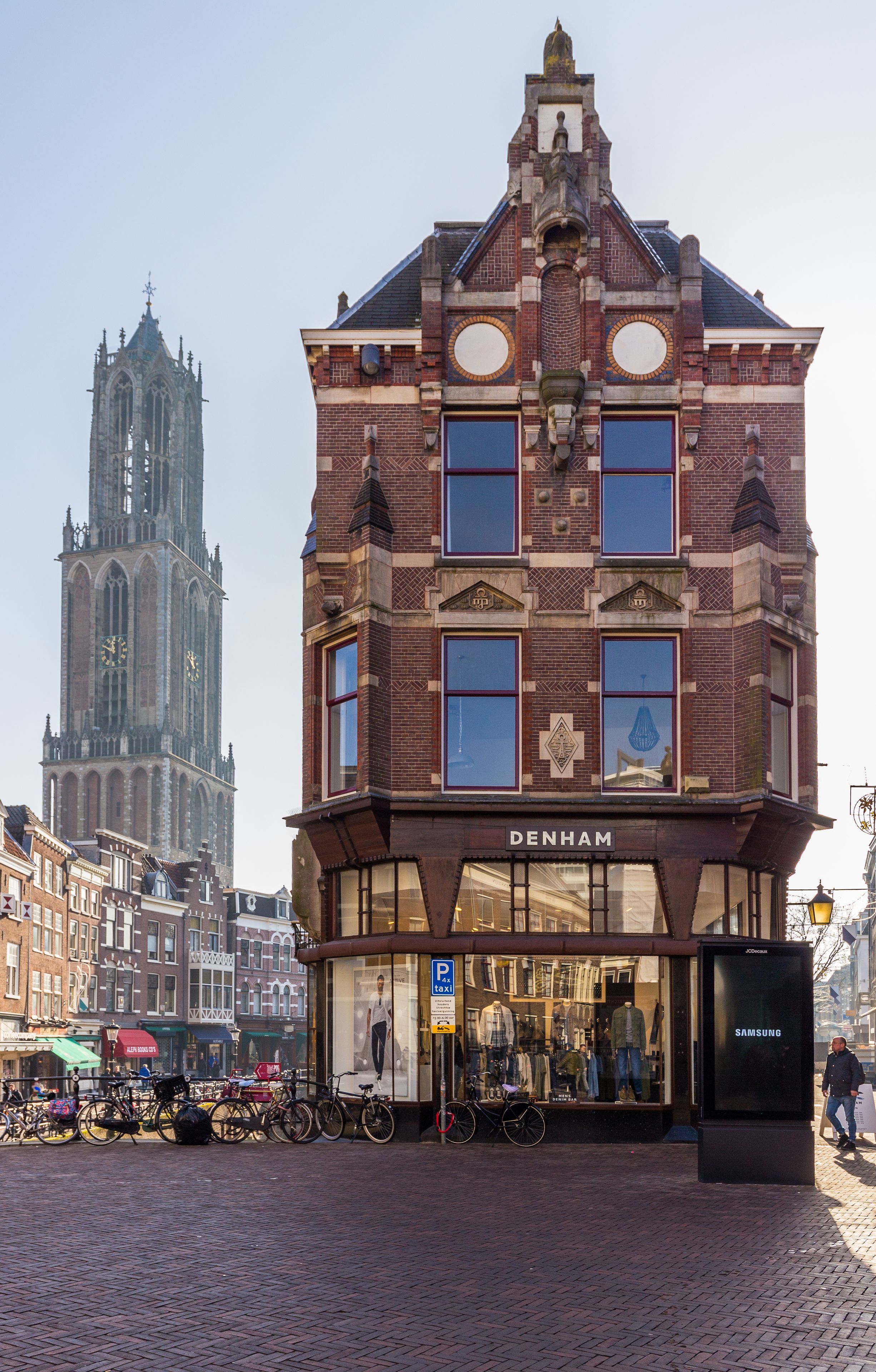 A historic building with a prominent clock tower in the background, a storefront with a sign reading "DENHAM," and bicycles parked outside.