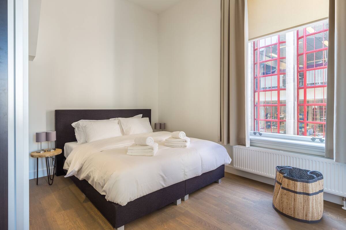 A neatly made bed with white linens and towels, a small round table with lamps, and a window with a red-framed view.