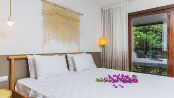 Una cama bien tendida con sábanas y almohadas blancas, un colgador de macramé en la pared, una lámpara de noche y vistas al verdor a través de una puerta de cristal.