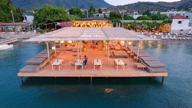 A floating restaurant with outdoor seating overlooking a body of water, surrounded by mountains and greenery.