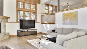 Ein geräumiges Wohnzimmer mit einem großen Flachbildfernseher, einem Holztisch, einem grauen Sofa und einem Kamin.