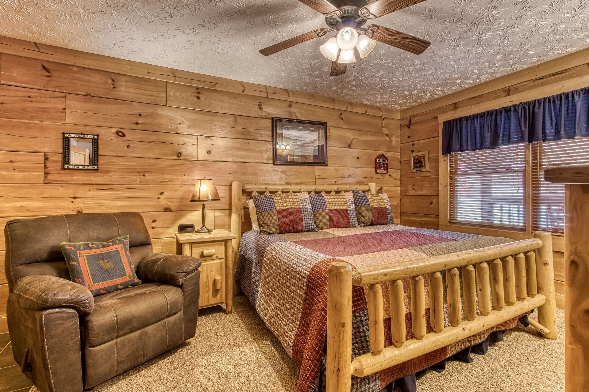 A cozy log cabin bedroom with a wooden bed, a leather armchair, a nightstand with a lamp, and a ceiling fan.