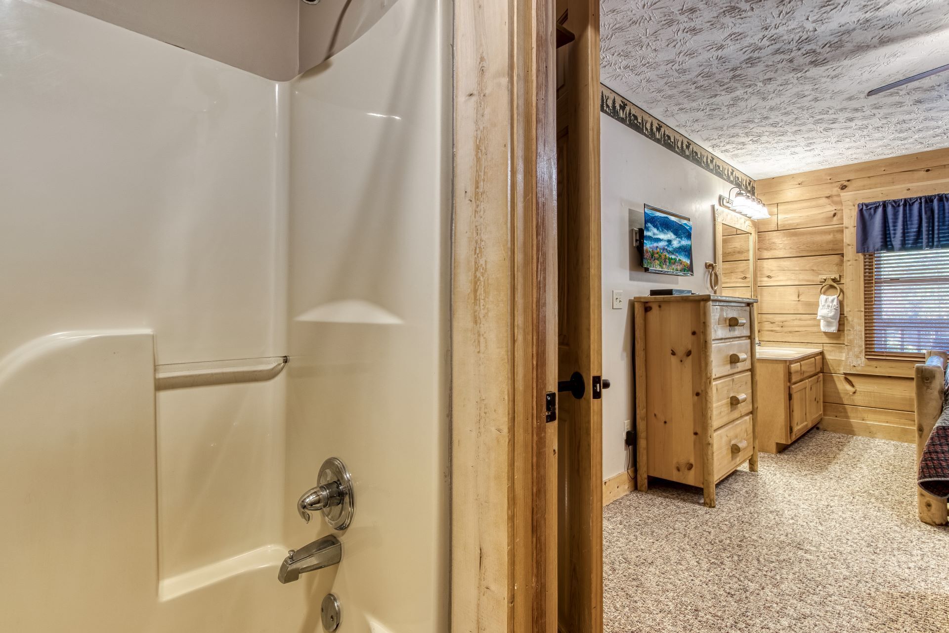 A wooden cabin interior with a white door, a wooden dresser, a small table with a towel, and a wall-mounted TV.