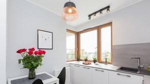 Una cocina moderna con armarios blancos, una silla negra, una mesa de comedor, un florero con flores rojas y un ventanal con vistas a edificios.