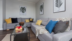 Una sala de estar moderna con dos sofás grises, cojines amarillos y azules, una mesa de centro de vidrio con manzanas, e impresiones enmarcadas de plantas en la pared.
