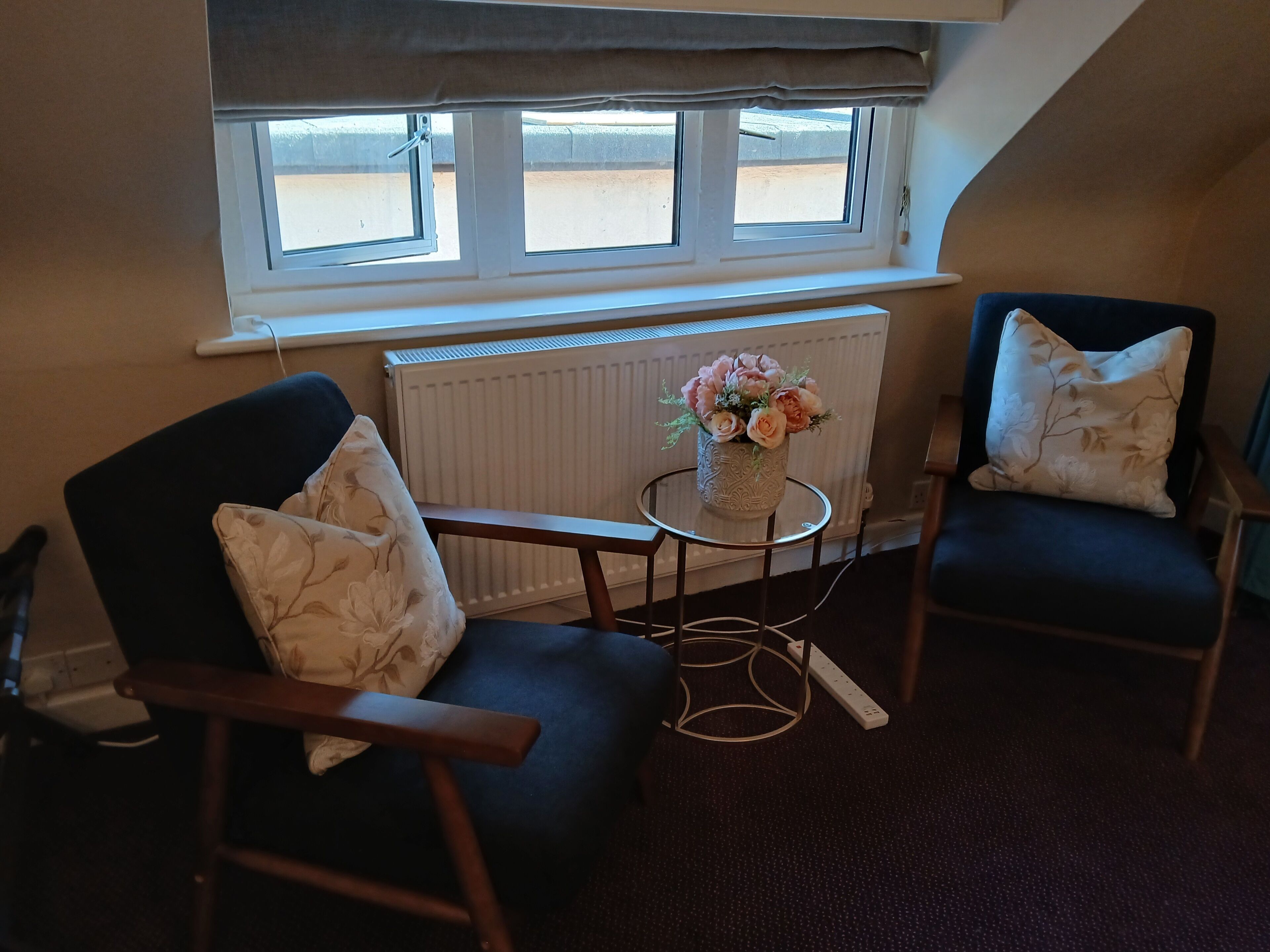 A room with two dark-colored armchairs, a small round table, and a vase with flowers. There is a window above the table and a radiator below.