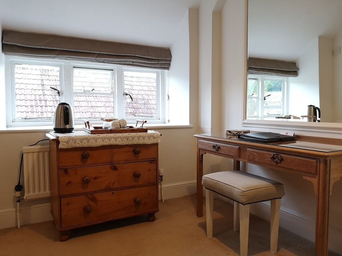 A room with a wooden dresser, a desk with a laptop, a kettle, and a window with a view of a brick building.