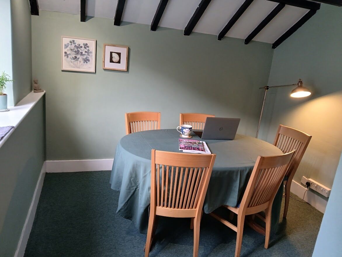 A room with a round table, four wooden chairs, a laptop, a lamp, and two framed pictures on the wall.