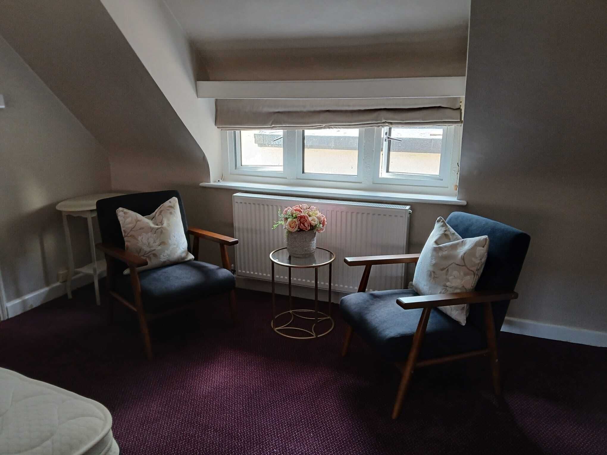A room with two chairs, a small table with a vase, and a window with blinds.