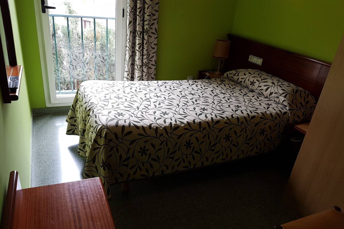 Une chambre d’hôtel avec un lit, une table de chevet, une lampe, une chaise en bois et une fenêtre donnant sur un paysage verdoyant.