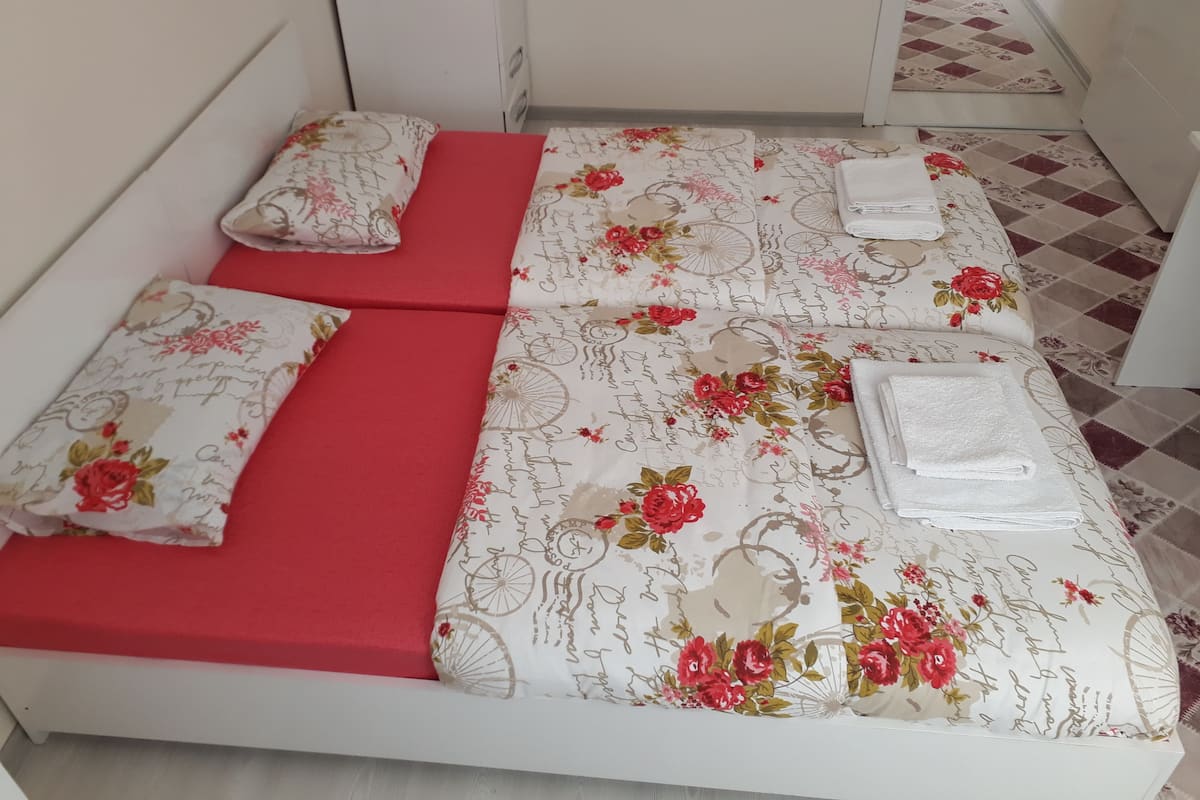 A small, neatly made bed with floral bedding and red accents, a white bedside table, and a checkered floor pattern.