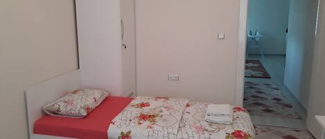 A single bed with a red mattress, a floral-patterned duvet, and a white pillow. A white wardrobe is positioned against the wall.