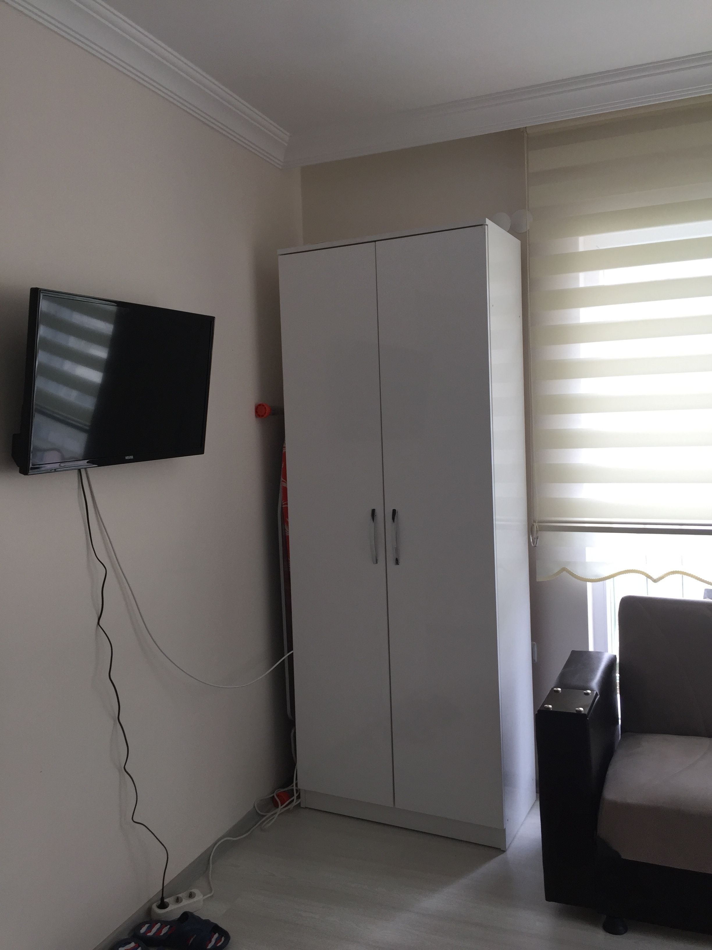 Una habitación con televisión montada en la pared, un armario, una silla y una ventana con persianas.