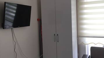 Una habitación con televisión montada en la pared, un armario, una silla y una ventana con persianas.