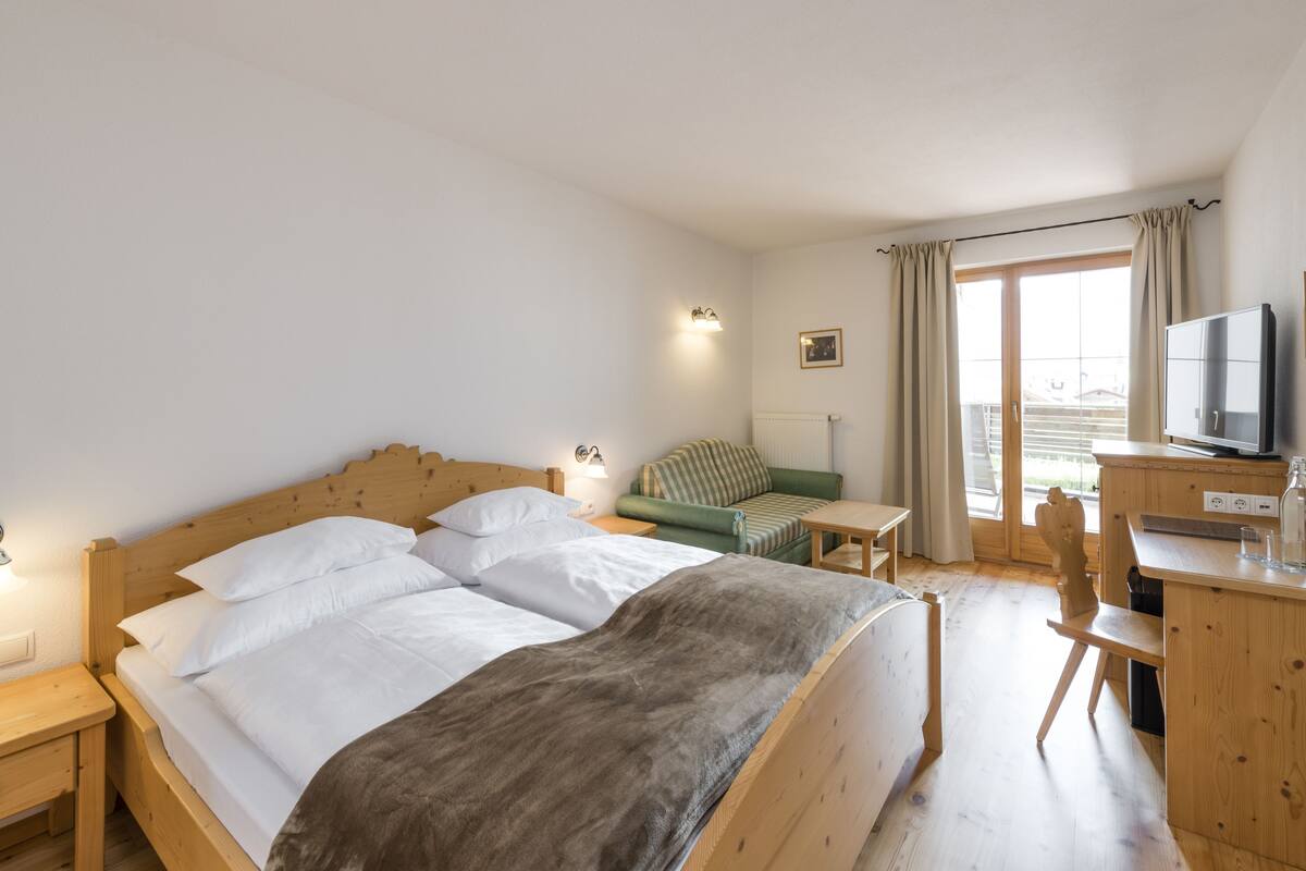 Une chambre d’hôtel avec un lit en bois, un petit bureau, une télévision et une fenêtre avec des rideaux.