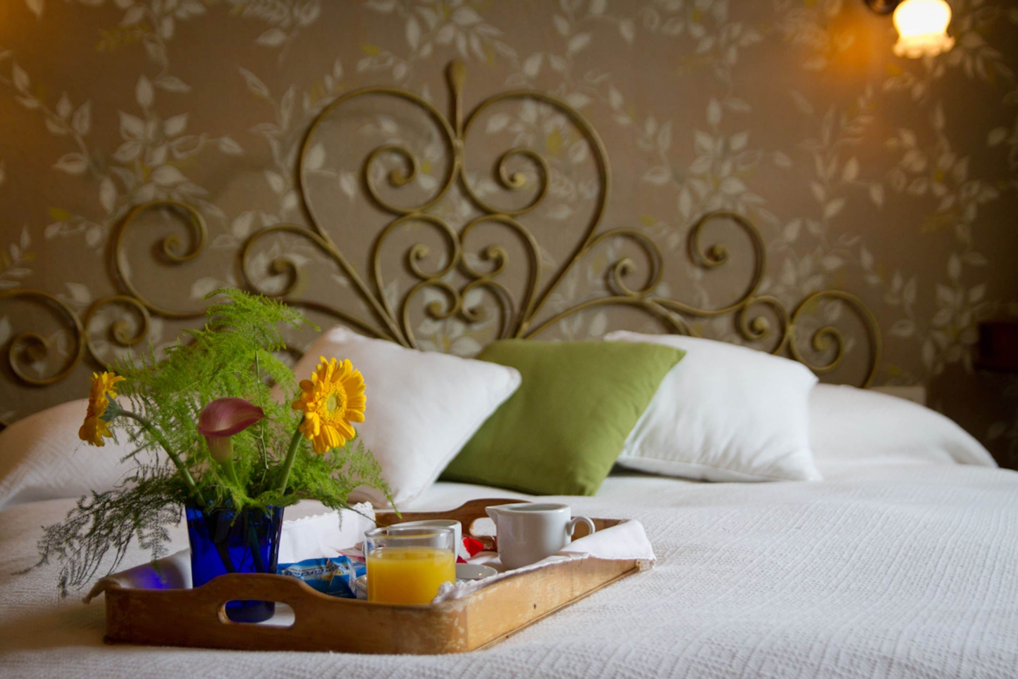 A neatly made bed with white bedding and a decorative headboard. A wooden tray with a blue vase, a glass of orange juice, and a cup is placed on the bed.