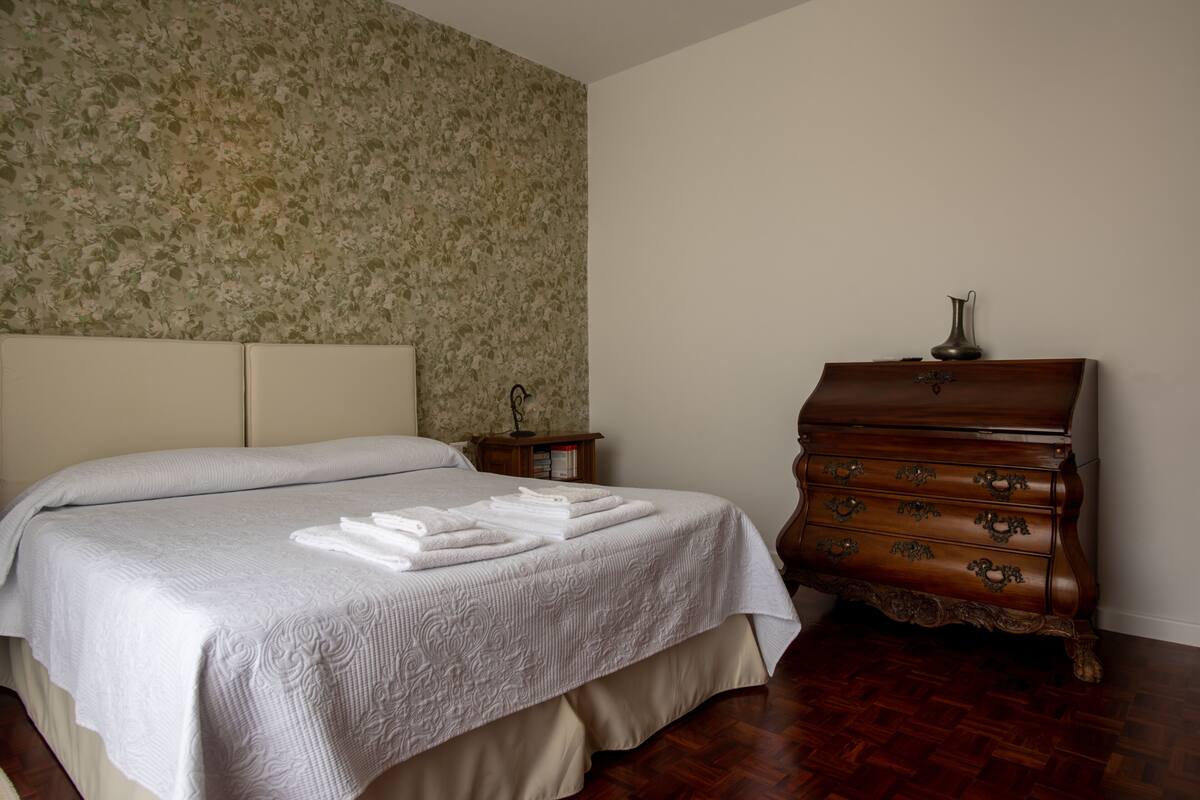 Una cama bien hecha con una colcha blanca y dos almohadas, un aparador de madera con cajones, y una mesita de noche con una lámpara y libros.