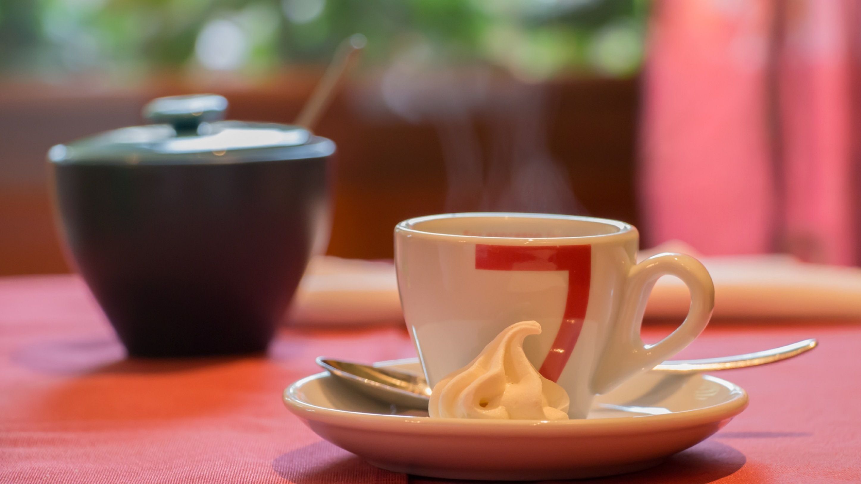 A cup of tea on a saucer with a spoon, a teapot, and a cup in the background.