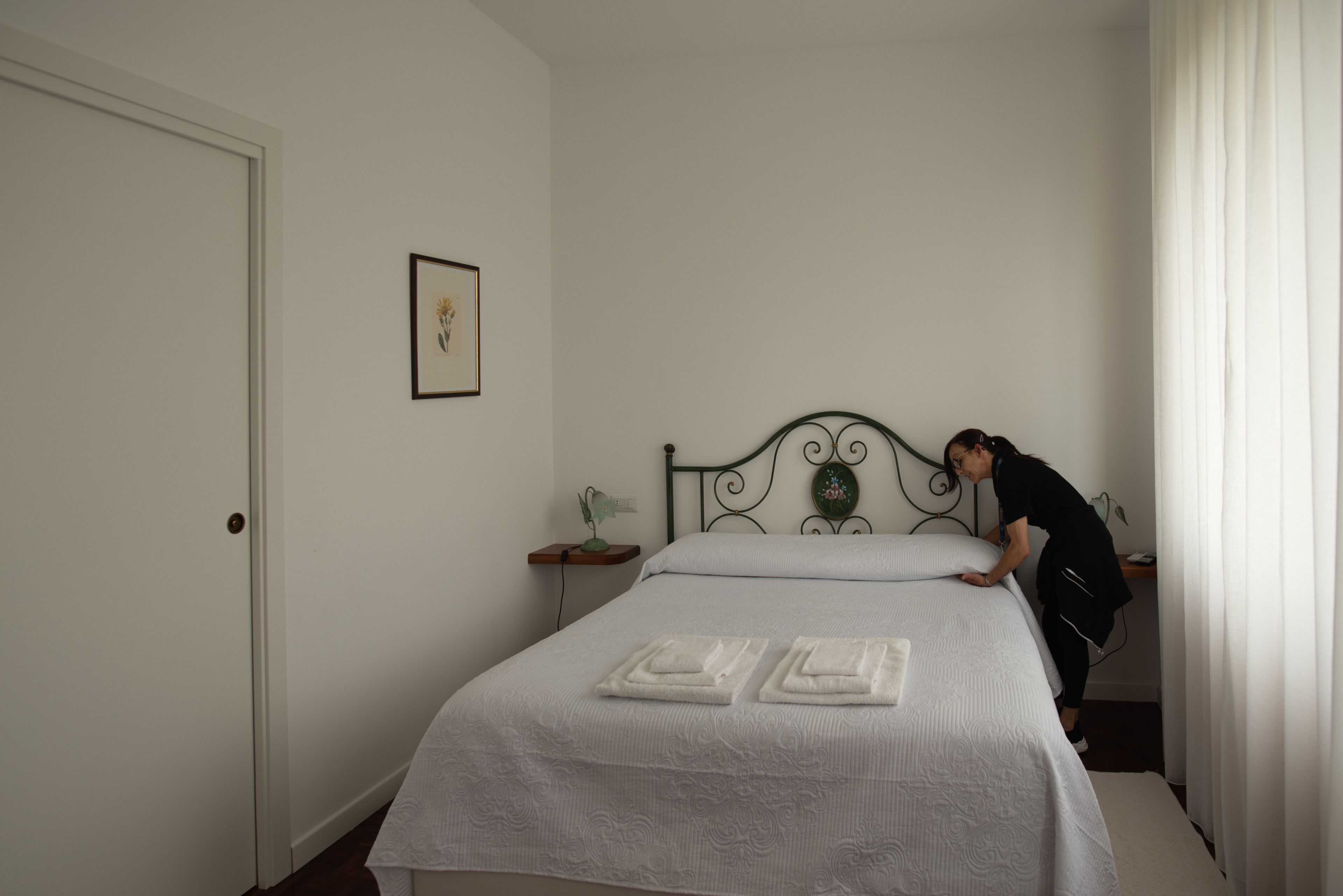 Cama bem arrumada com duas toalhas dobradas, mesa de cabeceira com abajur e um quadro na parede.