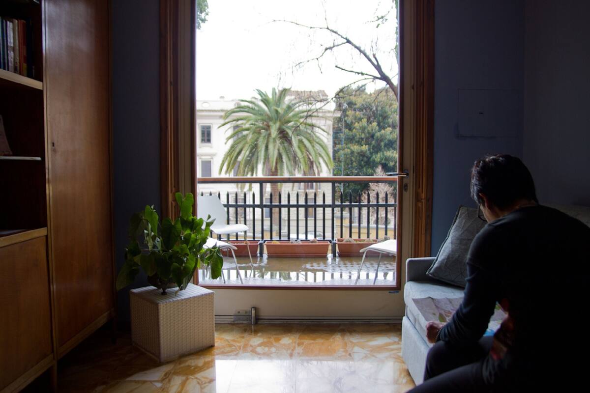 Una habitación con un ventanal grande, un balcón con barandilla, una planta en maceta y un sofá.