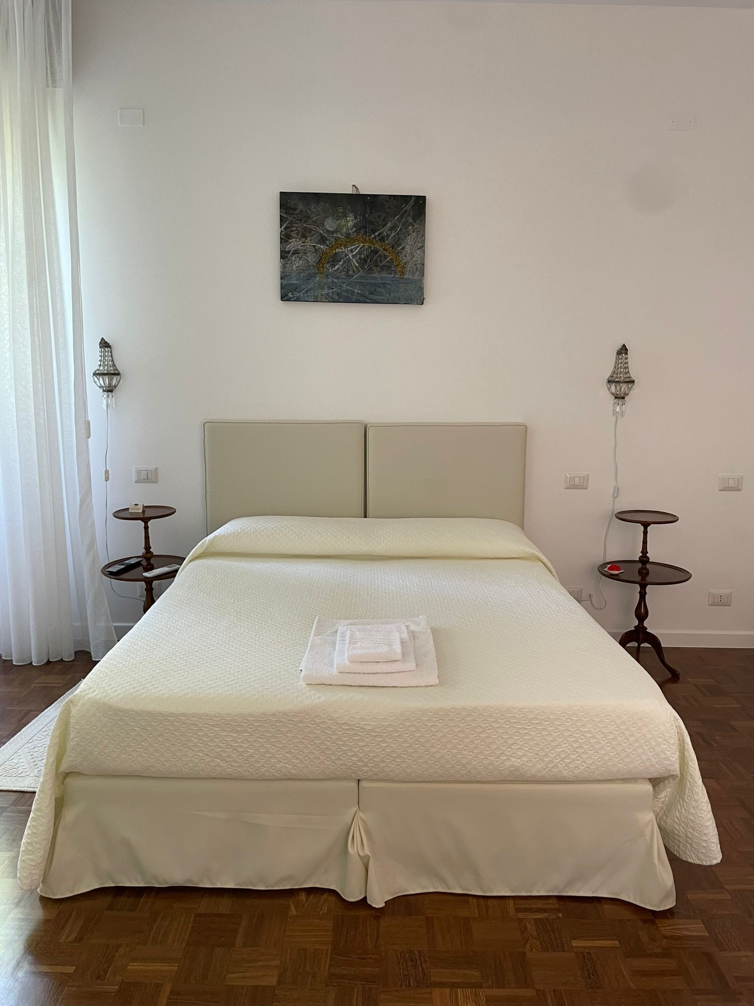 A neatly made bed with a white bedspread and a folded towel on it, two bedside tables with lamps, a painting on the wall, and a window with curtains.