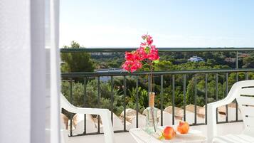 A balcony with white chairs, a table with fruits and a vase of flowers, and a view of greenery and buildings.