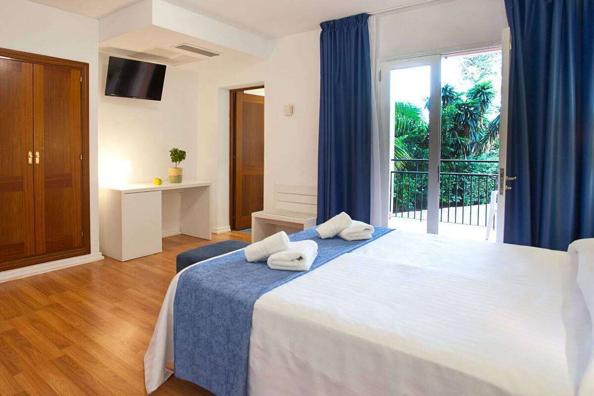 Ein Hotelzimmer mit einem Bett, einem Schreibtisch mit Stuhl, einem Fernseher und einem Balkon mit Vorhängen.
