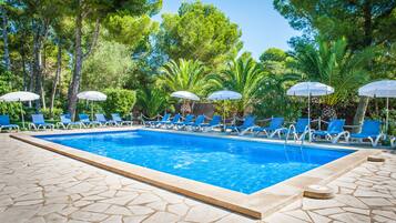 A swimming pool with blue water, surrounded by lounge chairs and umbrellas, set against a backdrop of trees and greenery.