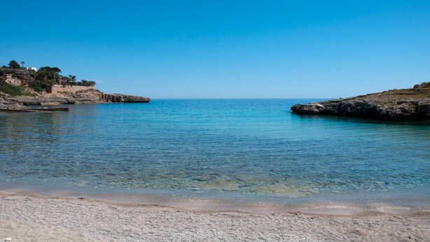 A coastal beach with clear water, rocky terrain, and a building on the left. - Hotel Cala Murada (Manacor)