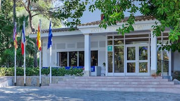 A hotel with a white facade, a tiled roof, and a sign that reads "HOTEL CALADA". There are flags in front of the hotel, and a stone wall with greenery around.