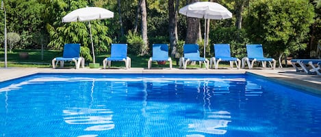 A swimming pool with blue water, surrounded by trees and blue lounge chairs with white umbrellas.