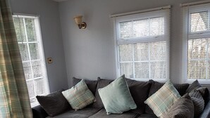 A sectional sofa with multiple cushions, a light-colored floor, and a window with blinds. - Tarrareoch Farm (Bathgate)