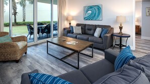 Una sala de estar moderna con un sofá gris, un sillón de mimbre y una mesa de centro. Hay un cuadro en la pared y una lámpara sobre una mesita auxiliar.