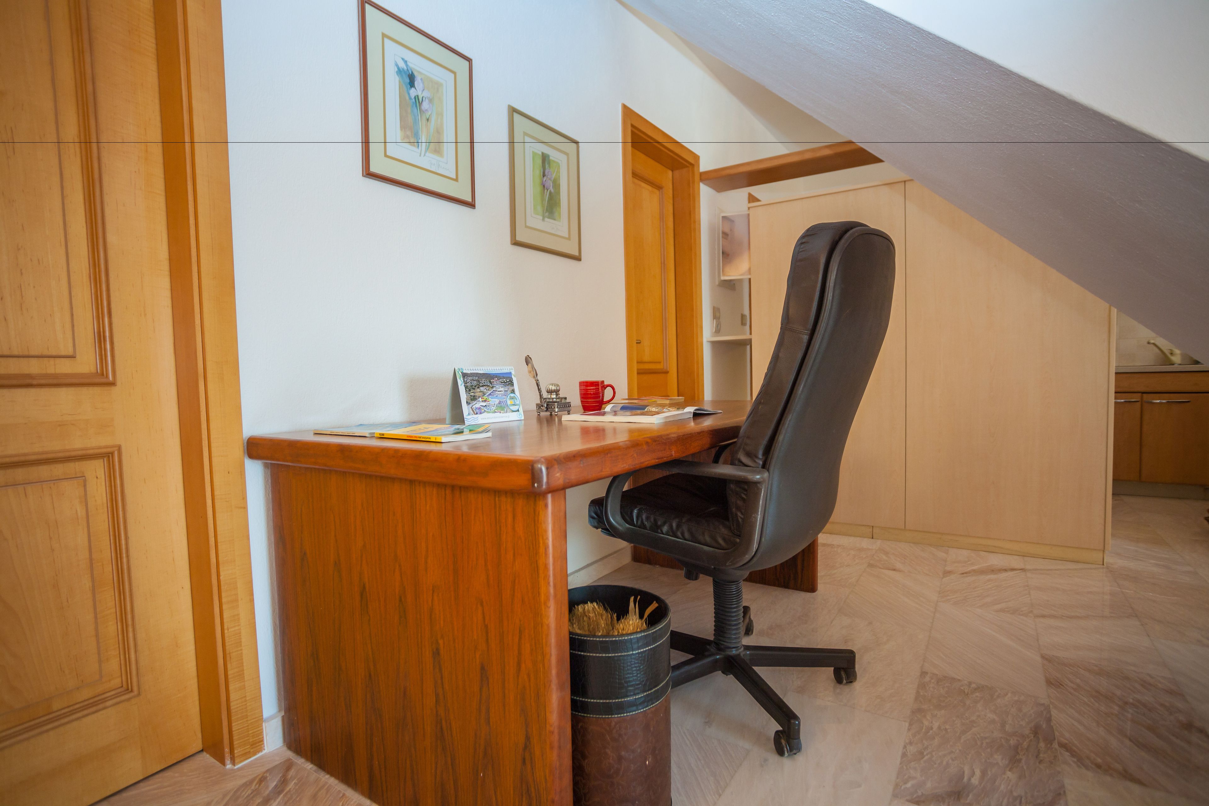 A small office space with a wooden desk, a black office chair, and a trash can. There are framed pictures on the wall and a door leading to another room.
