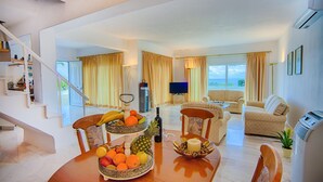 Ampla sala de estar com uma mesa de jantar posta com frutas, um sofá e uma televisão.