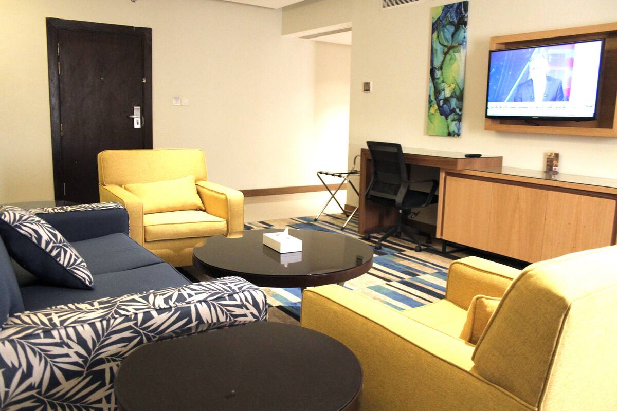 A hotel room with a sofa, armchair, and a small table. There is a TV mounted on the wall and a desk with a chair.