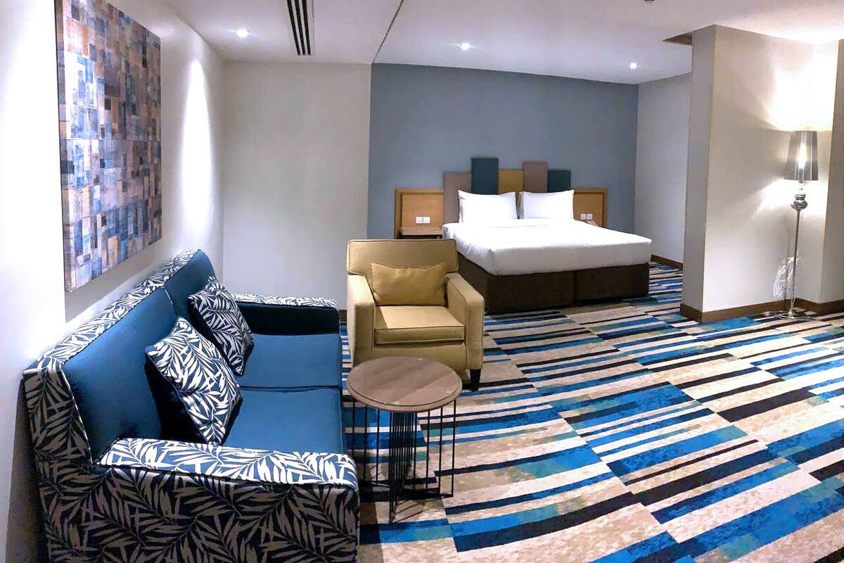 A hotel room with a blue sofa, a yellow armchair, a small round table, a bed, and a patterned rug.