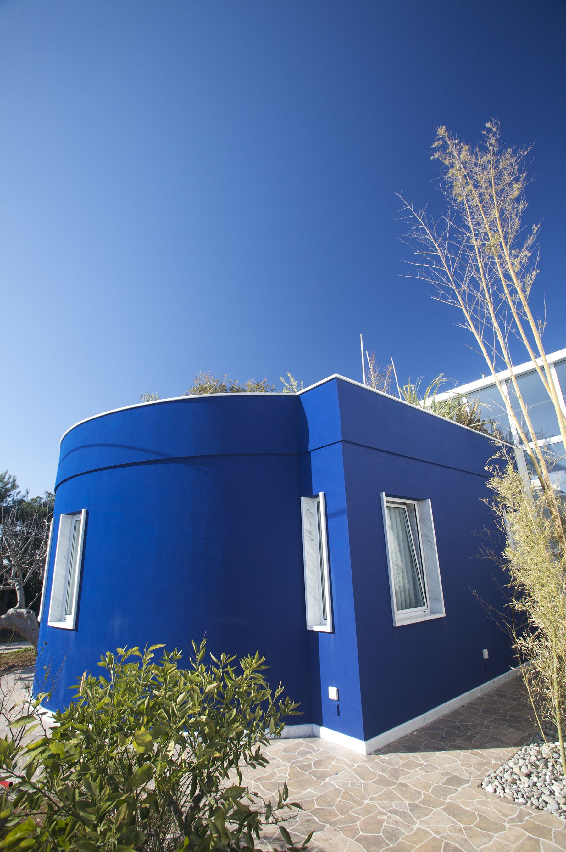 A modern blue building with a curved facade, large windows, and a stone-tiled entrance.
