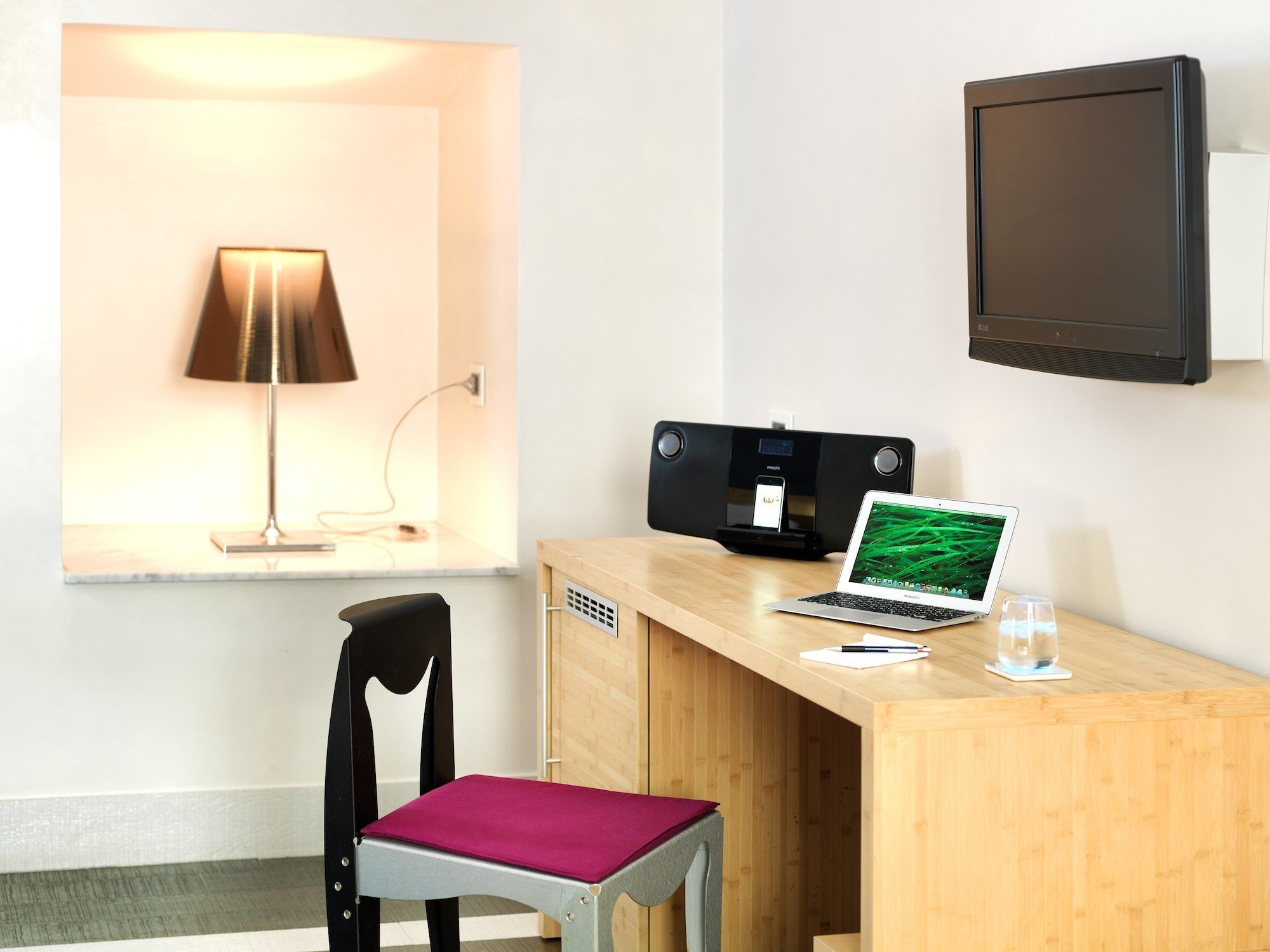 A hotel room with a wooden desk, a laptop, a smartphone on a charging station, a chair with a red cushion, a lamp, and a TV mounted on the wall.