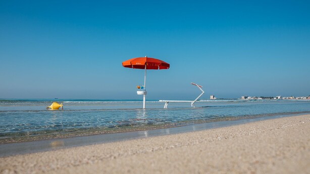 A beach with a red umbrella, a lifeguard tower, and a clear blue sky. - Hotel Intervallo (Porto Cesareo)