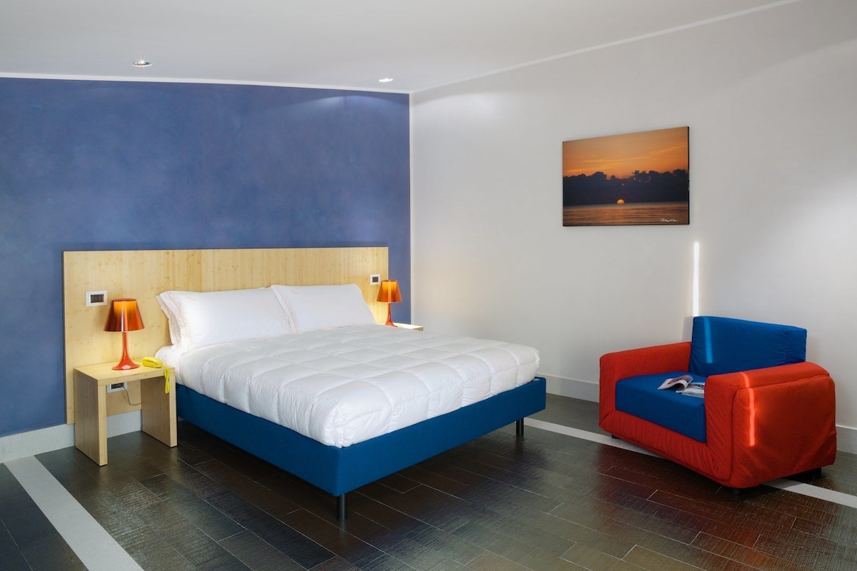 Una habitación de hotel moderna con una cama grande, una silla roja y azul, y un cuadro en la pared.